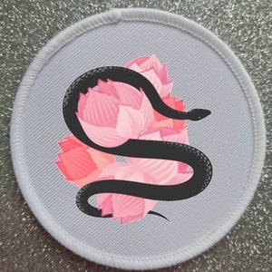 May include: A white circular patch with a black snake wrapped around pink lotus flowers. The snake is curled up and the flowers are in bloom.