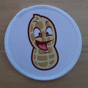 Può includere: Un cerotto rotondo bianco con un arachide di cartone animato con un viso sorridente e occhi marroni. L'arachide è marrone e giallo con una bocca e una lingua rosse.