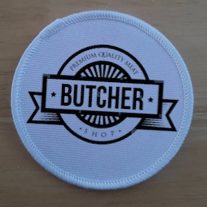 Op de afbeelding: Ronde witte stoffen patch met zwarte tekst die "Premium Quality Meat Butcher Shop" luidt.