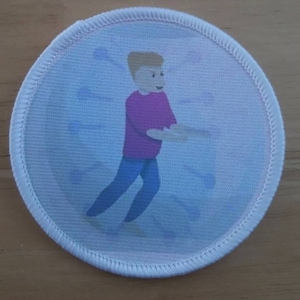 May include: A white round patch with a cartoon illustration of a person wearing a pink shirt and blue jeans. The person is holding a white object in their hands.