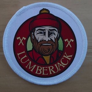 Peut inclure: Un écusson rond blanc avec un cercle rouge au centre. Le cercle présente une illustration de dessin animé d'un bûcheron avec un chapeau rouge et jaune, une barbe brune et un fond vert. Le texte "LUMBERJACK" est écrit en arc de cercle autour de l'image. Deux haches croisées se trouvent de chaque côté du bûcheron.
