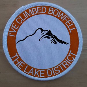 May include: A white and orange round patch with the text "I've Climbed Bowfell The Lake District" and a black silhouette of a mountain.