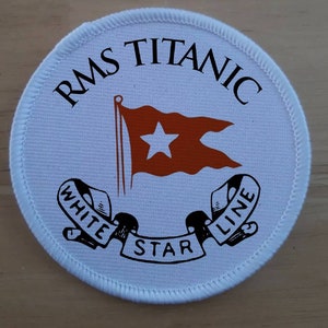 Op de afbeelding: Witte ronde patch met een rood-witte vlag, zwarte tekst met de tekst "RMS Titanic White Star Line" en een zwart lint met de woorden "White Star Line" in een cirkel.