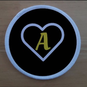 Puede incluir: Un parche circular blanco con fondo negro y un corazón blanco con una letra A dorada en el centro.