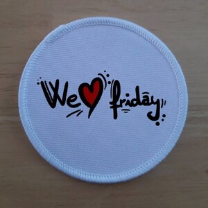 May include: White round fabric patch with black stitching around the edge. The patch has a red heart with the text "We ♥ Friday!" in black.