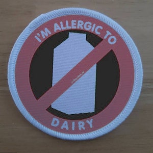 Può includere: Un cerotto rotondo bianco con un cerchio rosso e una linea rossa diagonale che attraversa un cartone di latte bianco. Il testo "I'm allergic to dairy" è scritto in bianco attorno al cerchio rosso.