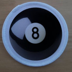 Puede incluir: Parche de tela blanco con un diseño de bola 8 en blanco y negro. La bola 8 está centrada en el parche y tiene un fondo blanco con un número 8 negro.