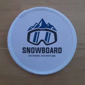 May include: White round patch with a blue and black snowboard goggle design and the text "SNOWBOARD EXTREME ADVENTURE".