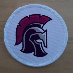 Puede incluir: Parche redondo blanco con un casco espartano morado y plateado con una pluma rosa.