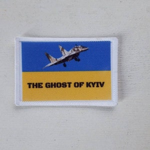 May include: A rectangular patch featuring the Ukrainian flag colors, blue and yellow, with a fighter jet graphic. The text "THE GHOST OF KYIV" is printed in black across the yellow section. The patch has a white border.