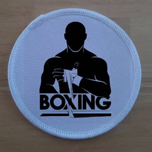 Può includere: Toppa rotonda bianca con una silhouette nera di un pugile che si avvolge le mani con la parola "BOXING" in lettere nere.