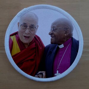 May include: A round white patch featuring a photograph of the Dalai Lama and Desmond Tutu smiling at each other. The Dalai Lama is wearing a red and yellow robe, while Tutu is wearing a black suit with a purple shirt and a silver chain with a cross pendant.