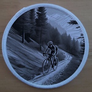 May include: Circular patch featuring a monochrome illustration of a cyclist on a road bike, navigating a forest path. The design includes a mountain scene and a white border.