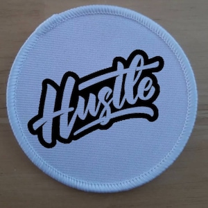 May include: White round fabric patch with a black outline and the word "Hustle" written in white cursive letters.