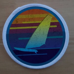 May include: A round white patch with a blue border. The center of the patch features a colorful sunset with a sailboat in the foreground. The sailboat is blue and white with a yellow sail.