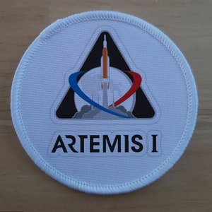 Könnte beinhalten: Ein weißer runder Patch mit einem schwarzen Dreieck, das eine weiße Rakete enthält, die von einem grauen Mond mit einer blauen und roten Umlaufbahn um ihn herum startet. Der Text "ARTEMIS I" ist in Schwarz unter dem Dreieck gedruckt.