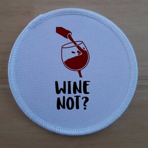 May include: White round patch with red wine being poured into a glass. The text "Wine Not?" is printed in black below the glass.