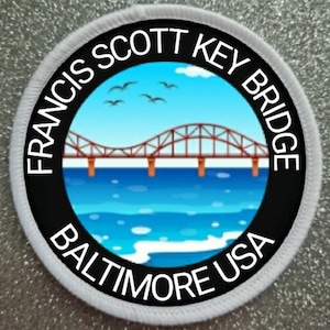 May include: A white and black circular patch with the text "Francis Scott Key Bridge Baltimore USA" around the edge. The center of the patch features a cartoon illustration of a red bridge over blue water with a blue sky and white clouds in the background.
