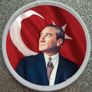 May include: A white circular patch with a red and white Turkish flag in the background. A man in a dark suit and tie is in the foreground, looking to the right.