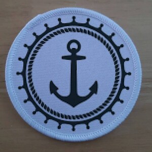 May include: White round fabric patch with a black anchor and a black rope design around the anchor. The anchor has a ring at the top and two points at the bottom.