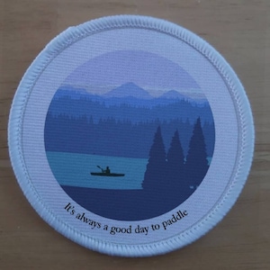 Könnte beinhalten: Runder weißer Patch mit einer blauen und violetten Landschaftsszene eines Sees, Bergen und Bäumen. Eine Silhouette einer Person in einem Kanu befindet sich auf dem See. Der Text "It's always a good day to paddle" befindet sich am unteren Rand des Patches.