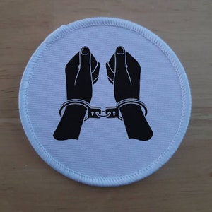 May include: White fabric patch with black silhouette of two hands cuffed together.