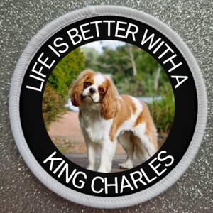 Op de afbeelding: Ronde patch met een witte rand en de tekst "LIFE IS BETTER WITH A KING CHARLES." De patch toont een Cavalier King Charles Spaniel hond in een natuurlijke omgeving. De hond heeft bruine en witte markeringen.