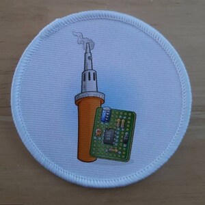 May include: A white round patch with a cartoon illustration of a soldering iron with smoke coming out of the tip and a circuit board with electronic components.