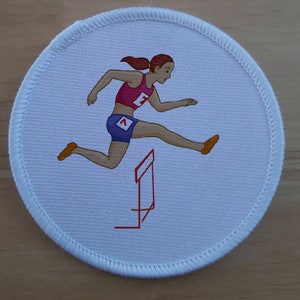 May include: A white round patch with a red border and a white stitched edge. The patch features a cartoon image of a female athlete wearing a pink tank top, blue shorts, and a red number "1" on her shirt. She is jumping over a red hurdle.