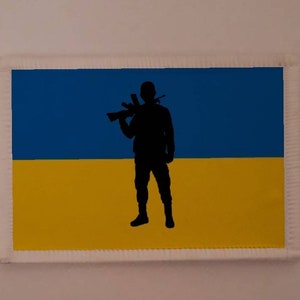 May include: A blue and yellow patch with a silhouette of a person holding a rifle. The patch is surrounded by a white border.
