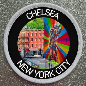 Op de afbeelding: Een witte en zwarte ronde patch met de tekst "Chelsea New York City" rond de rand. Het midden van de patch toont een kleurrijk muurschildering met een regenboogontwerp en een gebouw met rode bakstenen.