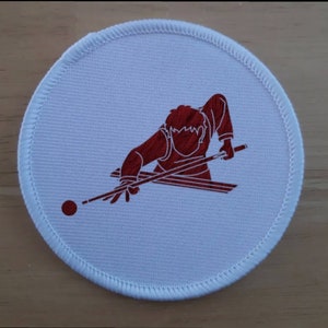 Puede incluir: Un parche redondo blanco bordado con una silueta roja de una persona jugando al billar. La persona sostiene un taco de billar y apunta a una bola roja.
