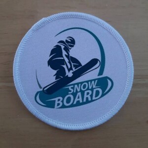 Op de afbeelding: Ronde witte patch met een blauwe en groene snowboarder-afbeelding en de tekst "SNOWBOARD" in een blauwe en groene boog.