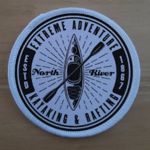 Puede incluir: Parche redondo blanco y negro con el texto "Extreme Adventure North River Kayaking & Rafting Est. 1867" y un gráfico de un kayak y remos.