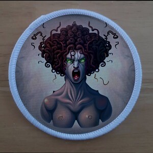 May include: A round white patch featuring a colourful illustration of Medusa, a mythical Greek figure with snakes for hair, a fierce expression, and green eyes.