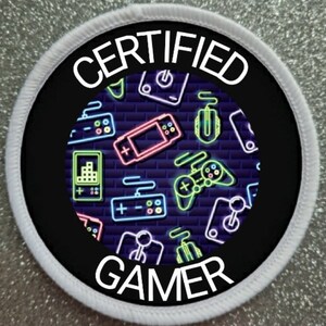 Peut inclure: Un patch rond blanc et noir avec le texte "CERTIFIED GAMER" en blanc. Le centre du patch présente un motif néon de manettes de jeu vidéo, de joysticks et d'autres icônes de jeu sur un fond bleu foncé.