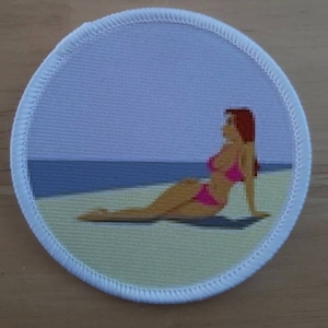 May include: A round white patch with a white border. The patch features a cartoon image of a woman in a pink bikini sitting on a beach with blue water in the background.