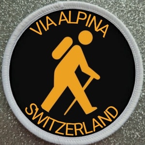 Op de afbeelding: Een witte ronde patch met een zwarte cirkel in het midden. De zwarte cirkel heeft de tekst "VIA ALPINA" in oranje letters rond de bovenrand en "SWITZERLAND" in oranje letters rond de onderrand. Een gele stickfiguur met een rugzak en een wandelstok bevindt zich in het midden van de zwarte cirkel.