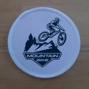 May include: White round patch with a black and white illustration of a mountain biker riding over a mountain. The text "MOUNTAIN BIKE" is printed in a black diamond shape.