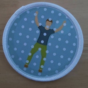 May include: A round, blue and white fabric patch with a white outline. The patch features a cartoon person with arms raised in the air, standing on a white polka dot background.