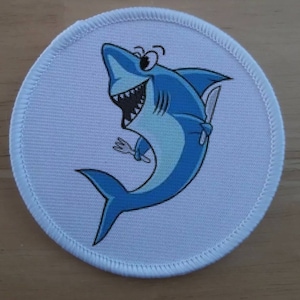 May include: A white round patch with a blue cartoon shark holding a fork and knife. The shark has a toothy grin and is facing the right side of the image.