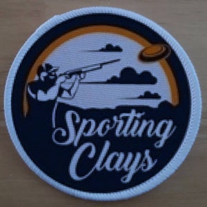 Puede incluir: Un parche circular blanco y naranja con fondo negro. El parche presenta la silueta de una persona disparando a una paloma de arcilla con el texto "Sporting Clays" en letras blancas en cursiva.