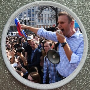 Peut inclure: Un homme portant une chemise à carreaux bleu et blanc parle dans un mégaphone tout en s'adressant à une foule de personnes. Il tient un drapeau russe dans sa main gauche et fait un geste de la main droite. La foule est rassemblée sur une place publique avec des bâtiments en arrière-plan.