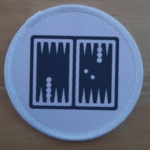 May include: A white round patch with a black outline of a backgammon board with black checkers.