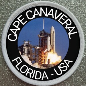 Op de afbeelding: Een ronde witte en zwarte patch met de tekst "CAPE CANAVERAL FLORIDA - USA" rond de rand. Het midden van de patch toont een kleurenfoto van een spaceshuttle die vanaf een lanceerplatform opstijgt.