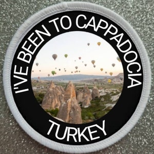以下が含まれることがあります： 黒い文字で「I've Been To Cappadocia Turkey」と書かれた白い丸いパッチと、岩の多い風景の上に熱気球が浮かんでいる写真です。
