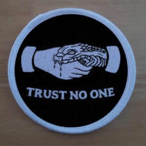 Könnte beinhalten: Runder, schwarzer Aufnäher mit weißem Rand. Das Design zeigt eine weiße Grafik eines Schlangenkopfes, der in eine Hand beißt, mit dem Text "TRUST NO ONE" in Weiß. Der Aufnäher ist wahrscheinlich zum Aufnähen oder Aufbügeln.