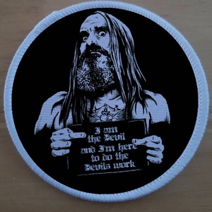 May include: A black and white embroidered patch featuring a man with long hair and a beard holding a sign that reads "I am the Devil and I'm here to do the Devil's work."