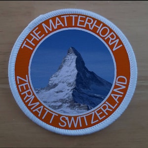 Könnte beinhalten: Ein runder weißer und orangefarbener Patch mit dem Text "THE MATTERHORN ZERMATT SWITZERLAND" am Rand. Die Mitte des Patches zeigt ein Foto des Matterhorns.