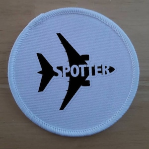 Peut inclure: Un patch rond blanc avec une silhouette d'avion noire et le mot "SPOTTER" imprimé dessus.